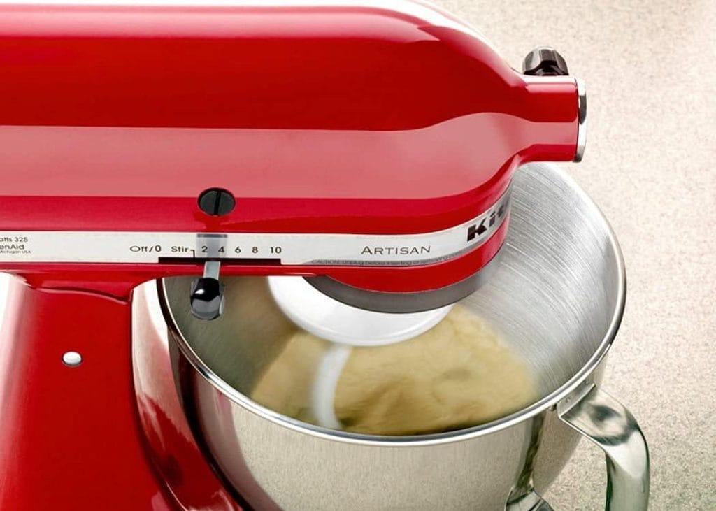 Close-up da batedeira KitchenAid Artisan Empire Red sovando massa clara em sua tigela de inox, com destaque para o seletor de 10 velocidades e o batedor gancho em movimento.