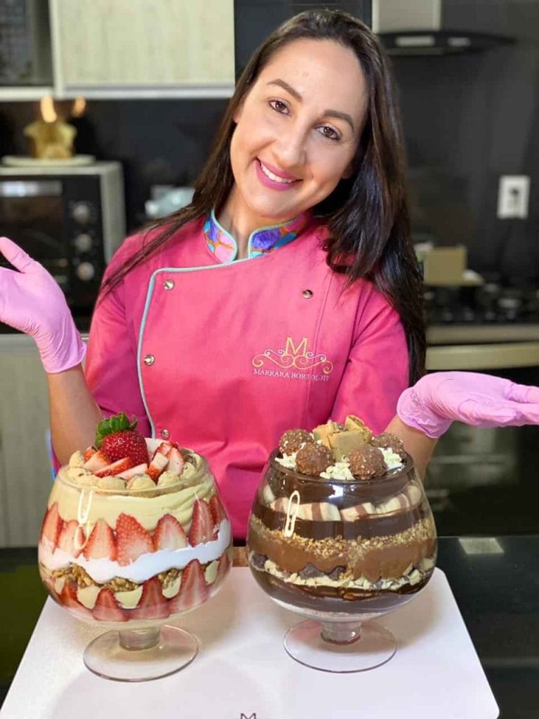Uma mulher sorridente em uma cozinha profissional, com duas grandes "Taças da Felicidade" prontas, uma de morango e outra de chocolate.