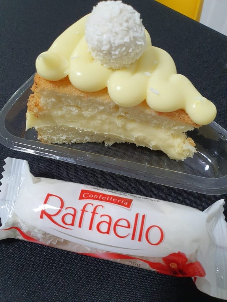 Uma fatia de bolo branco com recheio cremoso, coberto por uma calda de creme e um doce de coco, ao lado de uma embalagem de Raffaello.
