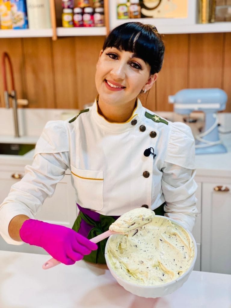 Uma mulher sorridente, vestindo um avental de confeiteira, segurando uma tigela grande com um recheio cremoso de chocolate e biscoito.