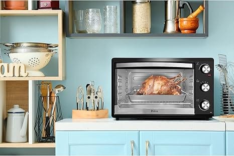 Forno elétrico Philco PFE44P de 44 litros, cor preta, com um frango assado dourado, em uma cozinha moderna com utensílios de confeitaria. Ideal para assar, tostar e gratinar receitas.