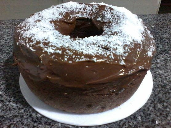 Um bolo de prestígio caseiro, com uma espessa cobertura escura de chocolate e uma generosa camada de coco ralado por cima, em um prato branco.