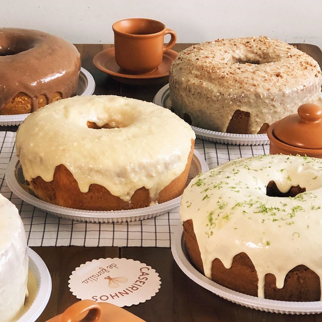 Quatro bolos caseiros redondos com coberturas de chocolate, glacê branco, coco ralado e limão fresco, dispostos em uma mesa com uma xícara.