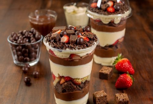 Copo da Felicidade gourmet em camadas com creme de Ninho, brigadeiro de chocolate, morangos frescos, pedaços de brownie e granulado. Foco no doce pronto para consumo, ao lado de morangos e brownies soltos. Ideal para confeiteiras e venda.