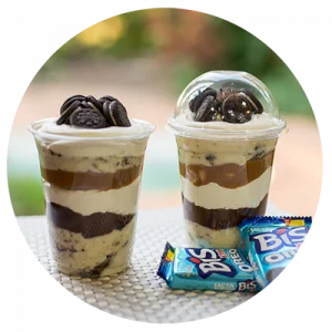Dois Copos da Felicidade com camadas de creme branco e biscoito Oreo triturado, cobertos com chantilly e biscoitos Oreo inteiros. Ilustra a variedade de sabores ensinados no curso Copos e Taças de Sucesso.