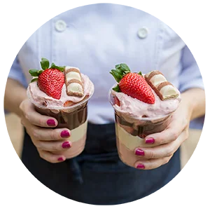 Chef Isis Alvarez mostrando Copos da Felicidade com camadas de frutas vermelhas, creme e pedaços de Kinder Bueno, decorados com morangos frescos. Exemplifica a criatividade e o apelo visual ensinados no curso.