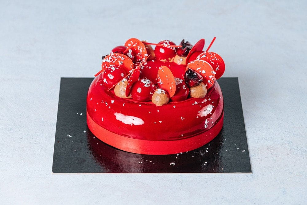 Um entremet com glacage espelhado vermelho vibrante, decorado com frutas vermelhas, raspas de chocolate e folhas de prata comestível.