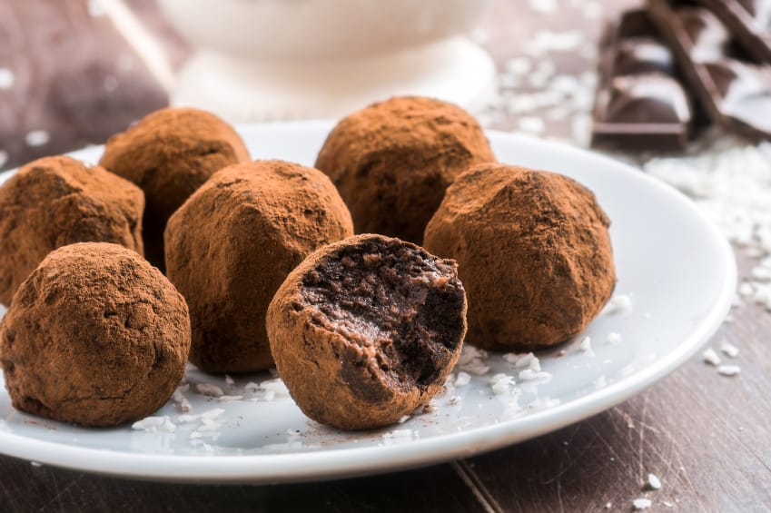 Trufas de chocolate clássicas, macias, com uma delas partida ao meio mostrando a textura do recheio, e cobertas com cacau em pó.