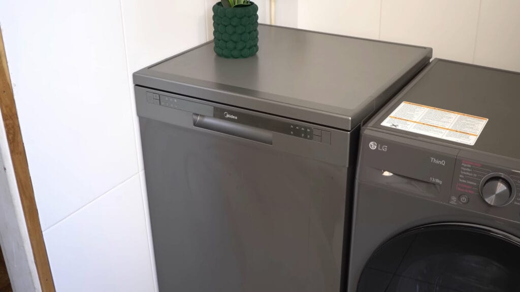 Lava-louças Midea 14 serviços preta instalada em uma cozinha profissional ao lado de uma máquina de lavar, destacando o acabamento polido e o painel digital superior.