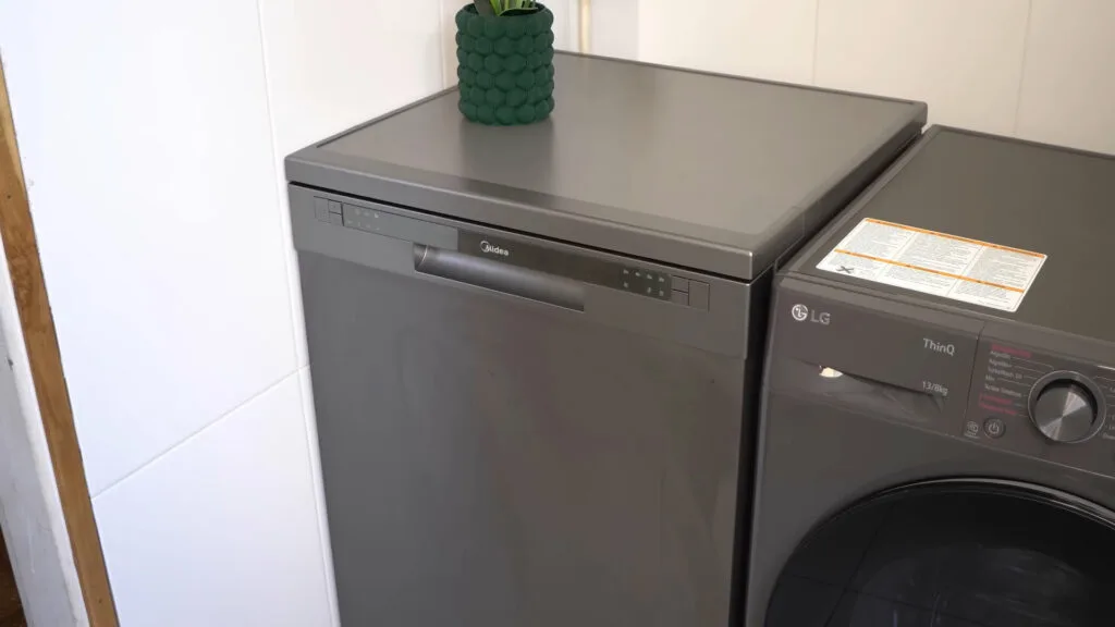 Lava-louças Midea 14 serviços preta instalada em uma cozinha profissional ao lado de uma máquina de lavar, destacando o acabamento polido e o painel digital superior.