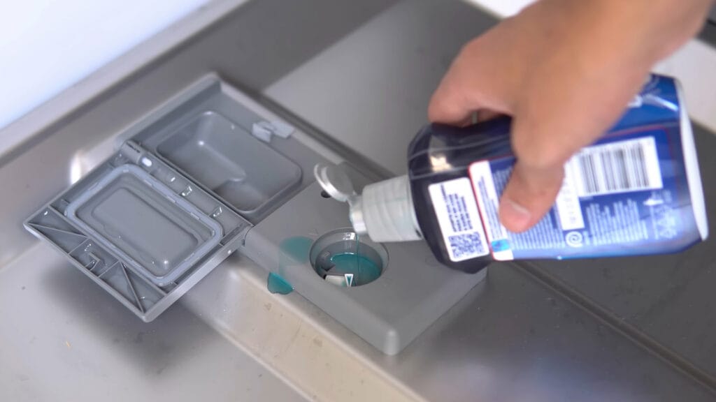 Mão abastecendo o reservatório de líquido secante em uma lava-louças Midea 14 serviços aberta, destacando a praticidade do compartimento na porta interna.