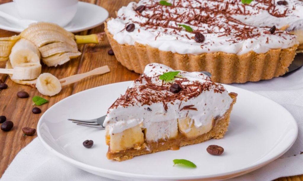 Torta banoffee com uma fatia cortada e servida em um prato branco, mostrando a base de biscoito, doce de leite, fatias de banana e chantilly com raspas de chocolate.