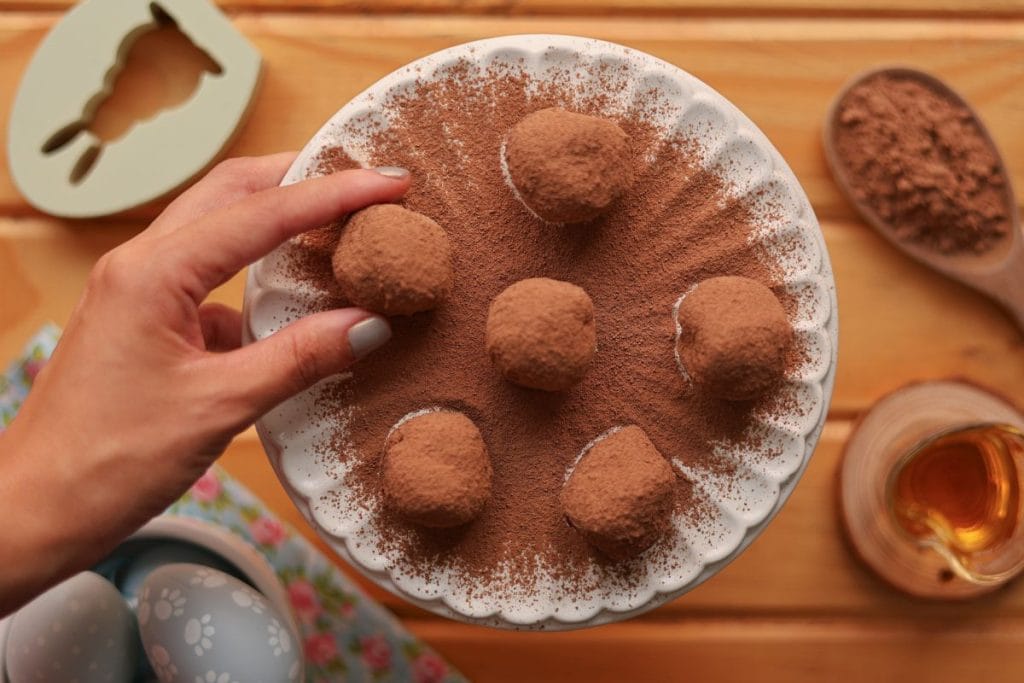 Mão pegando uma trufa de chocolate coberta com cacau em pó de um prato branco.