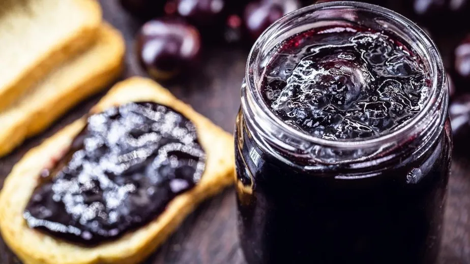 Close-up de um pote de geleia de uva caseira escura, com pedaços de fruta, ao lado de uma fatia de pão torrado com geleia, evidenciando a textura e o brilho. Uvas frescas e torradas desfocadas ao fundo.