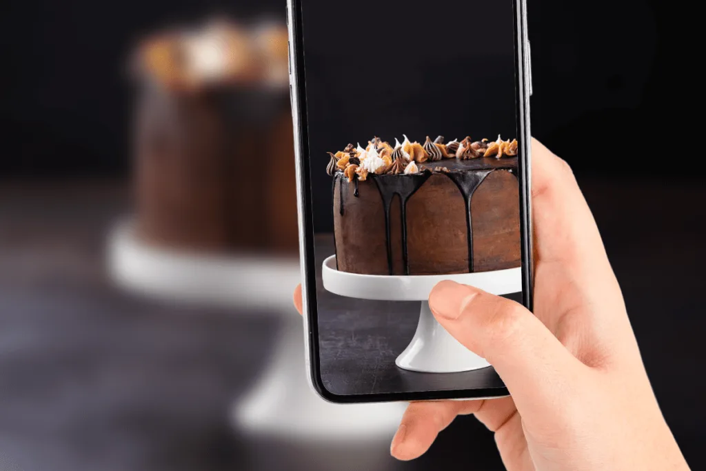 Uma mão segurando um smartphone tirando uma foto de um bolo de chocolate escuro e decorado com cobertura escorrendo e doces no topo, em um fundo escuro.
