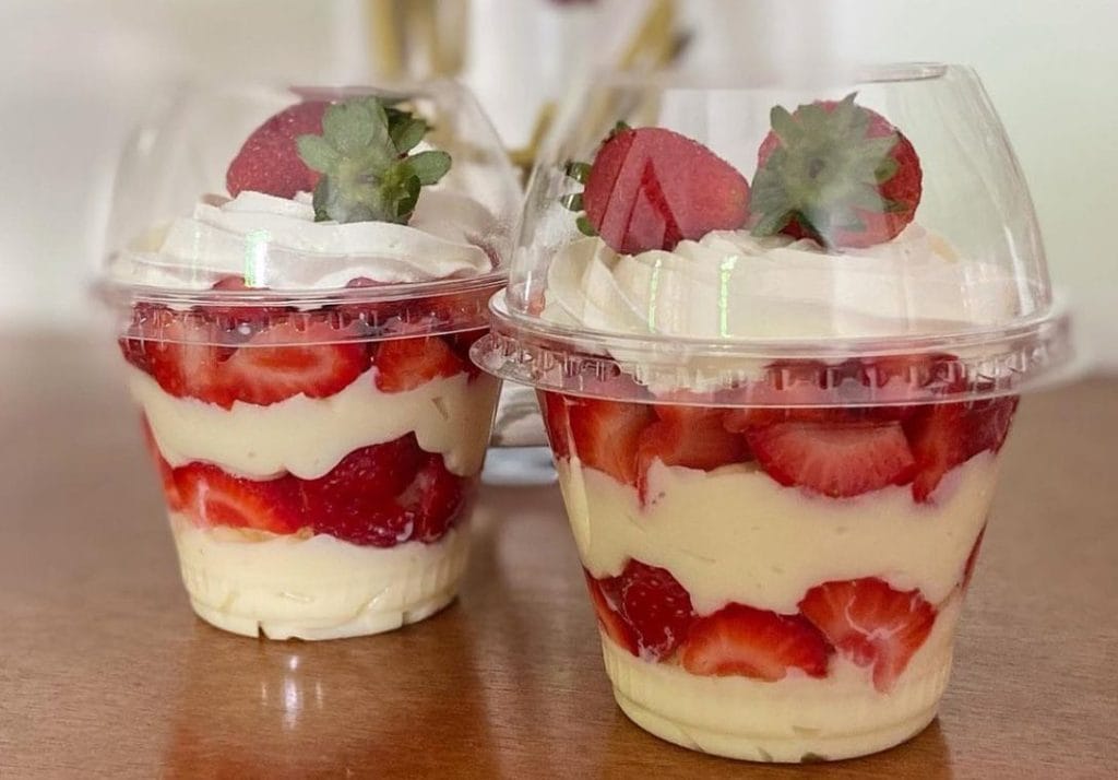 Dois Copos da Felicidade com camadas visíveis de creme de Ninho, morangos frescos fatiados e chantilly, decorados com morangos inteiros. Ideal para o tema de confeitaria, sobremesas e vendas.