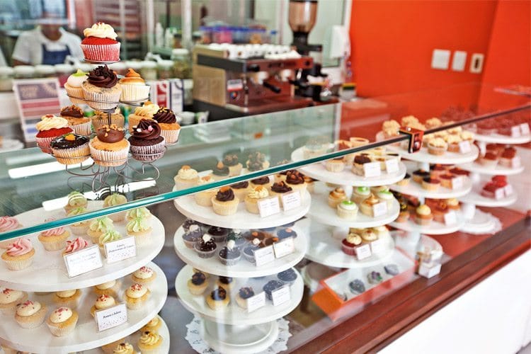Uma vitrine de vidro de uma confeitaria moderna, exibindo uma variedade de cupcakes coloridos em suportes, fatias de tortas decoradas e outros doces finos em exposição.