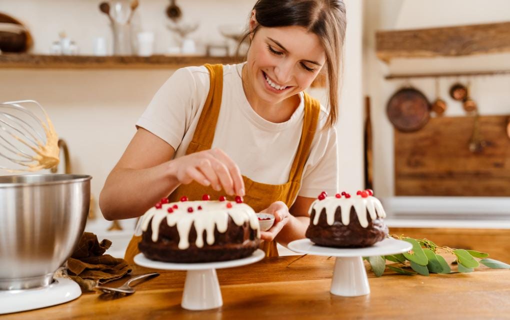 Mulher sorrindo em uma cozinha profissional, decorando um bolo com frutas vermelhas, representando a paixão e o crescimento da pequena confeitaria.