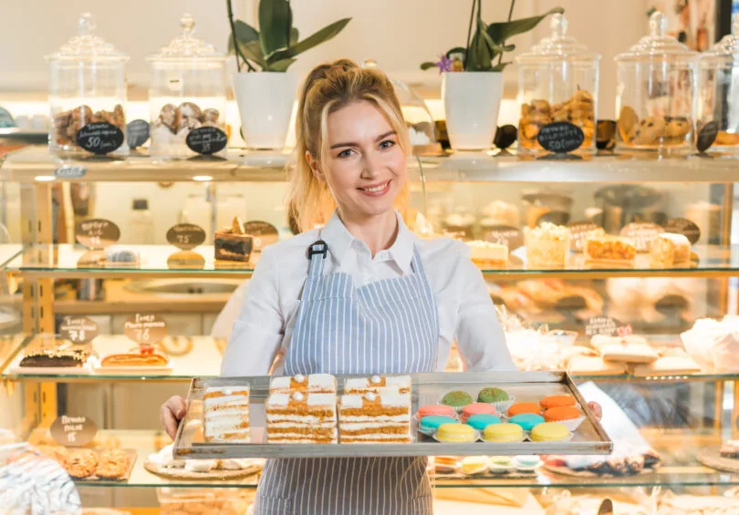 Uma confeiteira sorridente vestindo avental, segurando uma bandeja com fatias de bolo e macarons, em uma loja com doces e bolos em vitrines de vidro ao fundo.