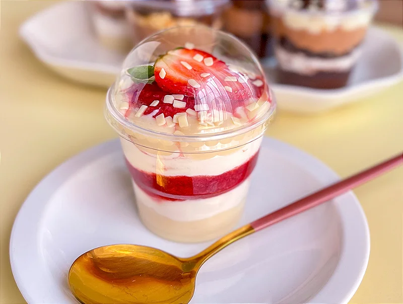 Copo da Felicidade em camadas com creme branco, calda de frutas vermelhas e pedaços de fruta, finalizado com chantilly, morango fresco fatiado e raspas de chocolate branco. Inclui colher dourada. Ideal para confeitaria, sobremesas leves e festas.