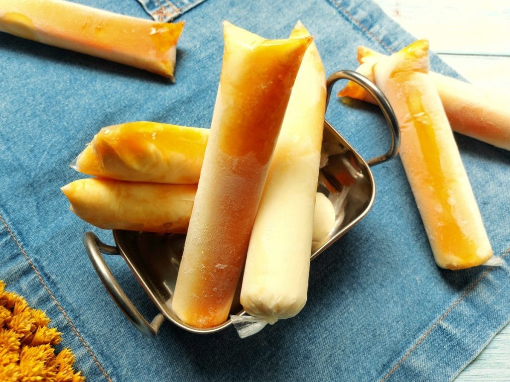 Close-up de dindins de pudim cremosos e alaranjados com calda de caramelo visível, dispostos em um recipiente prateado sobre fundo de tecido jeans.
