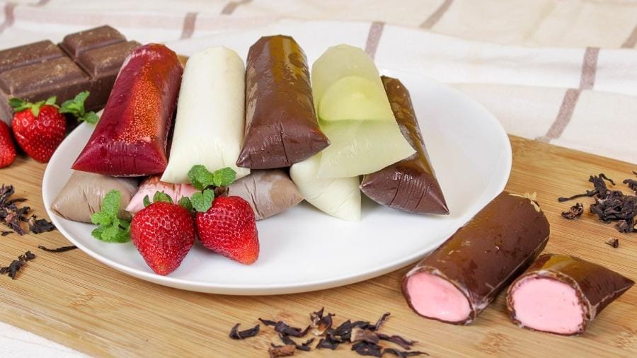 Plateau com diversos dindins gourmet coloridos, morangos frescos e barras de chocolate em um fundo de madeira com pano de prato. Dois dindins de chocolate com recheio rosa na frente.