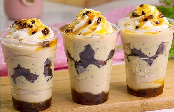 Três Copos da Felicidade com camadas de mousse de maracujá, pedaços de brownie escuro e calda de maracujá, decorados com chantilly e sementes de maracujá. Imagem para confeitaria, doces gourmet e venda.