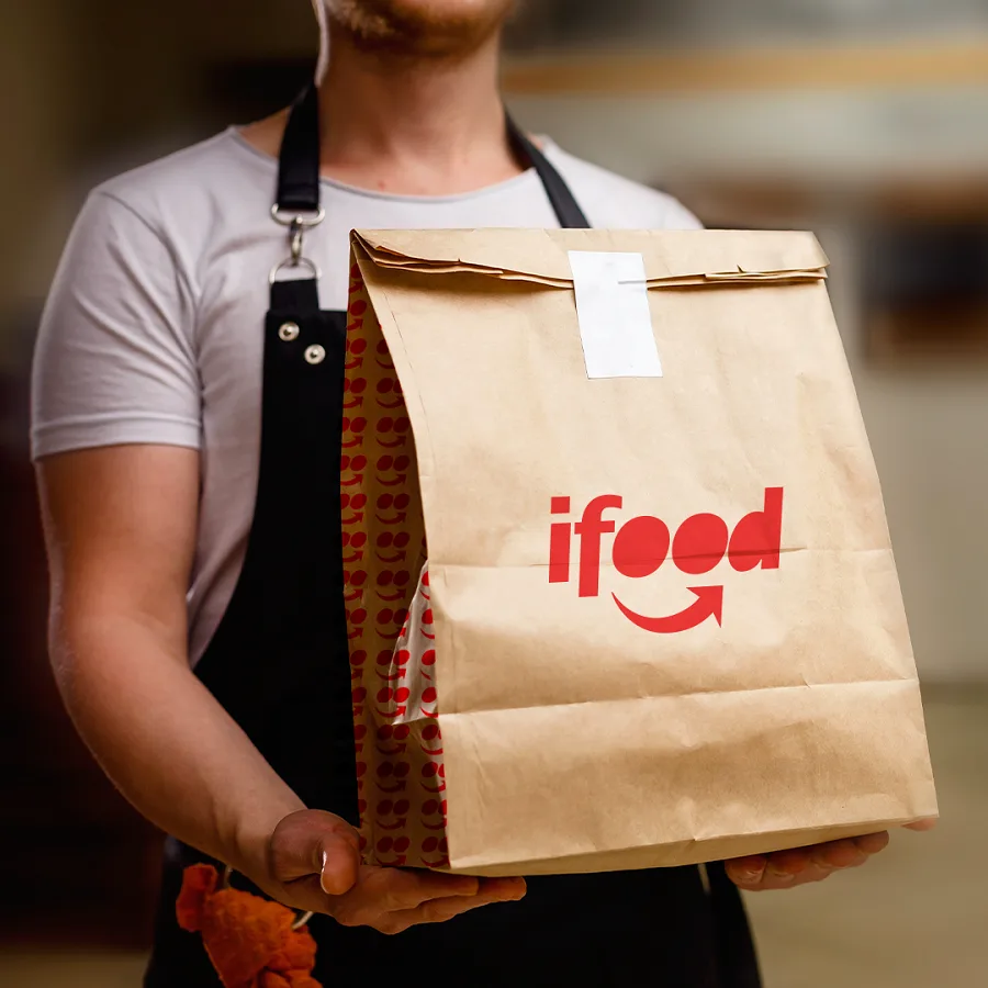 Um empreendedor de gastronomia vestindo avental, segurando uma sacola de papel kraft da iFood pronta para entrega, simbolizando o serviço de delivery.
