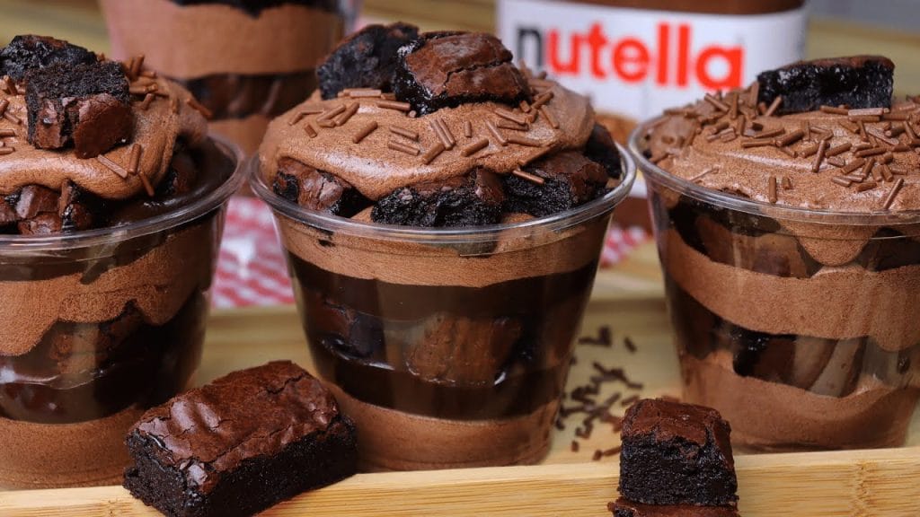 Copo da Felicidade com camadas de creme de chocolate, pedaços de brownie e granulado, decorado com mais brownie e um pote de Nutella ao fundo. Perfeito para confeitaria, sobremesas de chocolate e venda.