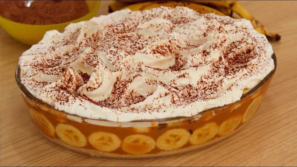 Banoffee de travessa de vidro com camadas de banana e doce de leite visíveis, coberta com chantilly e polvilhada com cacau em pó.