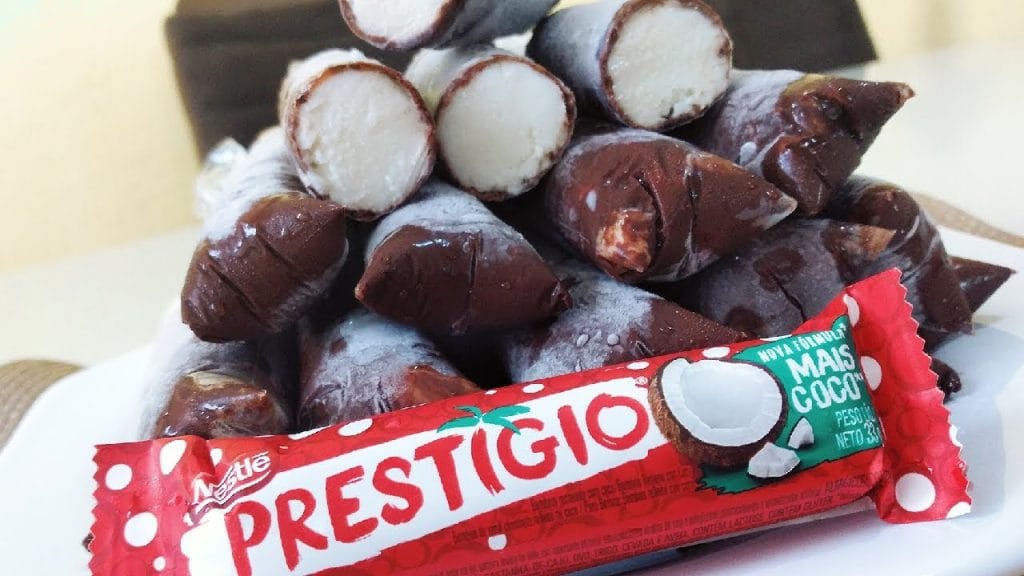 Pilha de dindins de Prestígio com recheio branco de coco e cobertura escura de chocolate, com uma barra de chocolate Prestígio em primeiro plano. Evidencia a inspiração no bombom.