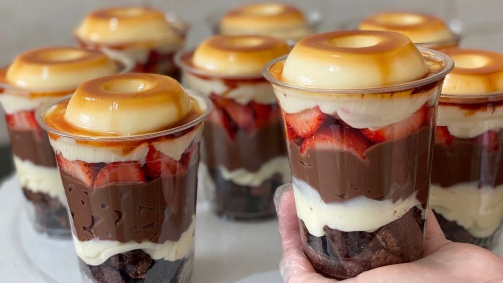 Muitos Copos da Felicidade com camadas de pudim, brigadeiro de chocolate, morangos frescos e creme branco, com calda de caramelo visível no topo do pudim. Ideal para receitas, confeitaria e venda.