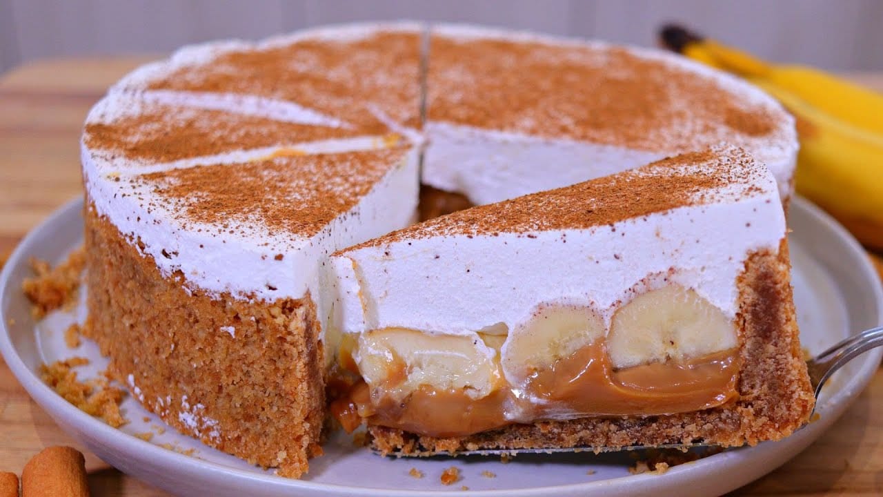 Torta banoffee com fatia cortada mostrando as camadas de biscoito, doce de leite e banana com chantilly e canela.