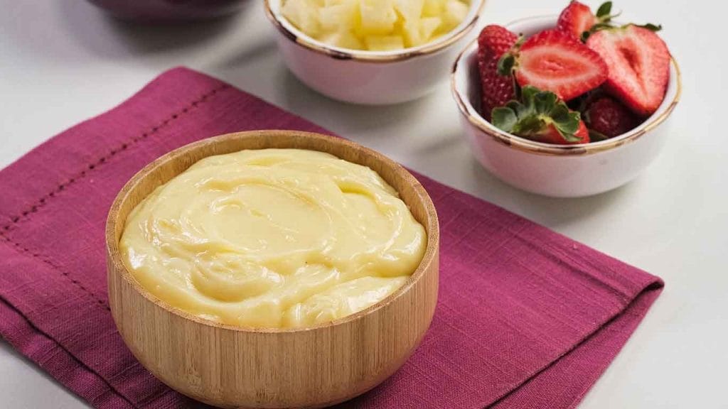 Uma tigela de madeira contendo recheio cremoso de leite ninho, com tigelas menores em segundo plano contendo morangos fatiados e pedaços de abacaxi.