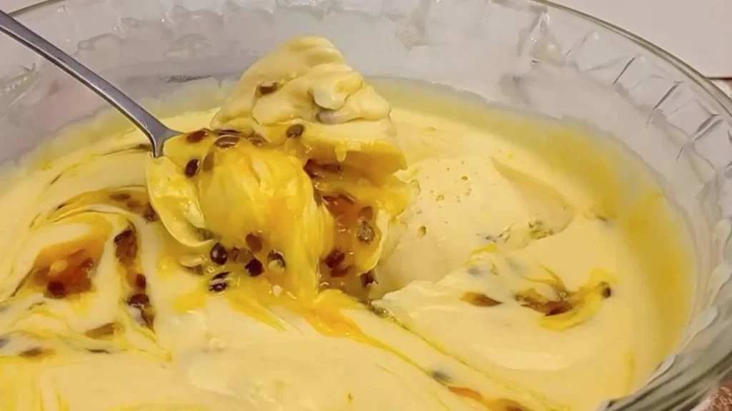 Uma colher de metal retirando recheio cremoso de leite ninho misturado com polpa e sementes de maracujá, em uma tigela de vidro.