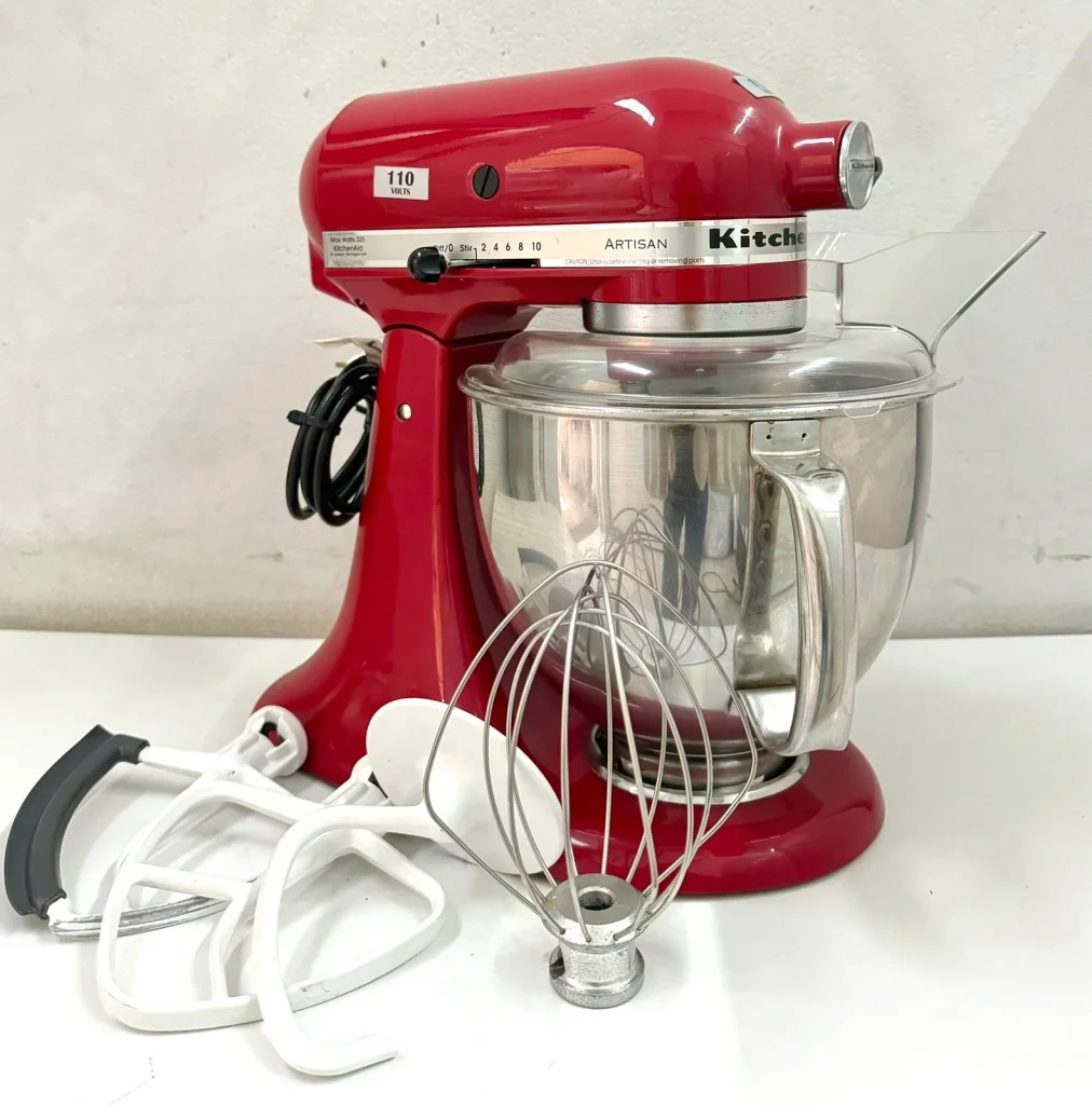 Conjunto completo da batedeira KitchenAid Artisan Empire Red sobre uma superfície branca, incluindo a tigela com alça, protetor de respingos, batedor fouet, pá plana e gancho para massas.