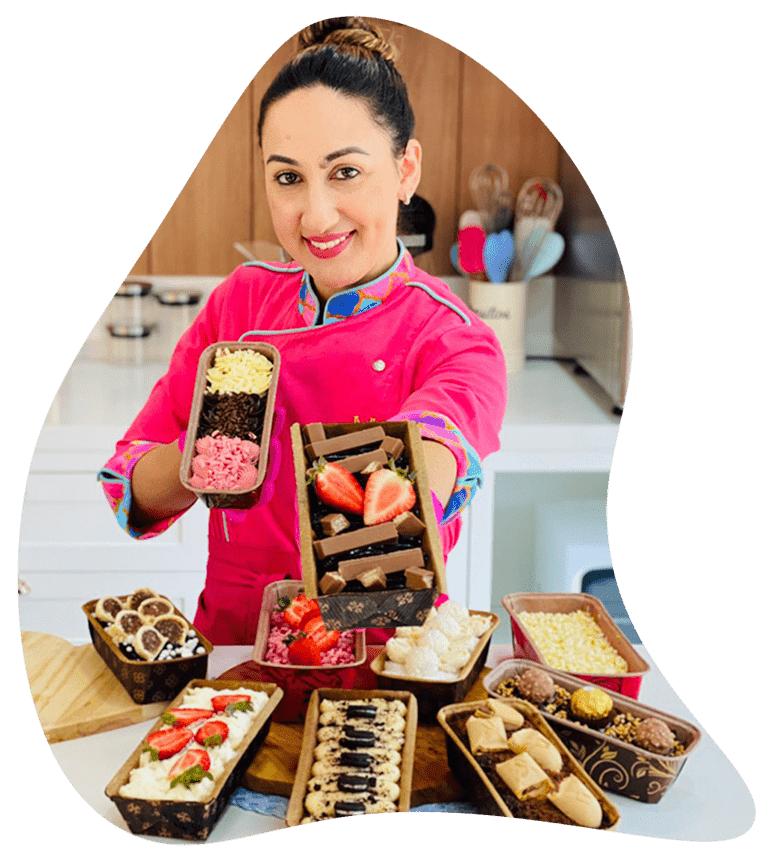 Chef confeiteira sorridente exibindo diversos mini bolos caseirinhos (slice cakes) e bolos de pote com cobertura e frutas, prontos para delivery e venda