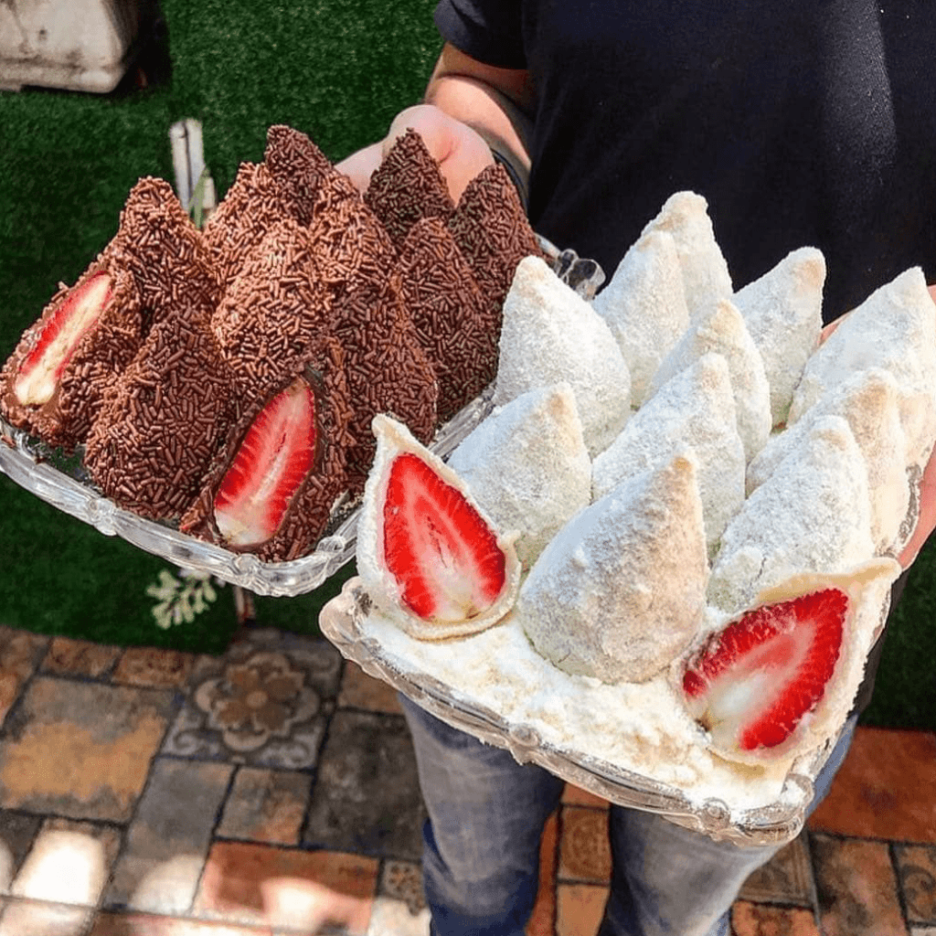 Pessoas segurando bandejas de trufas ou morangos trufados com casca de chocolate e granulado de chocolate ou leite em pó, com morangos frescos