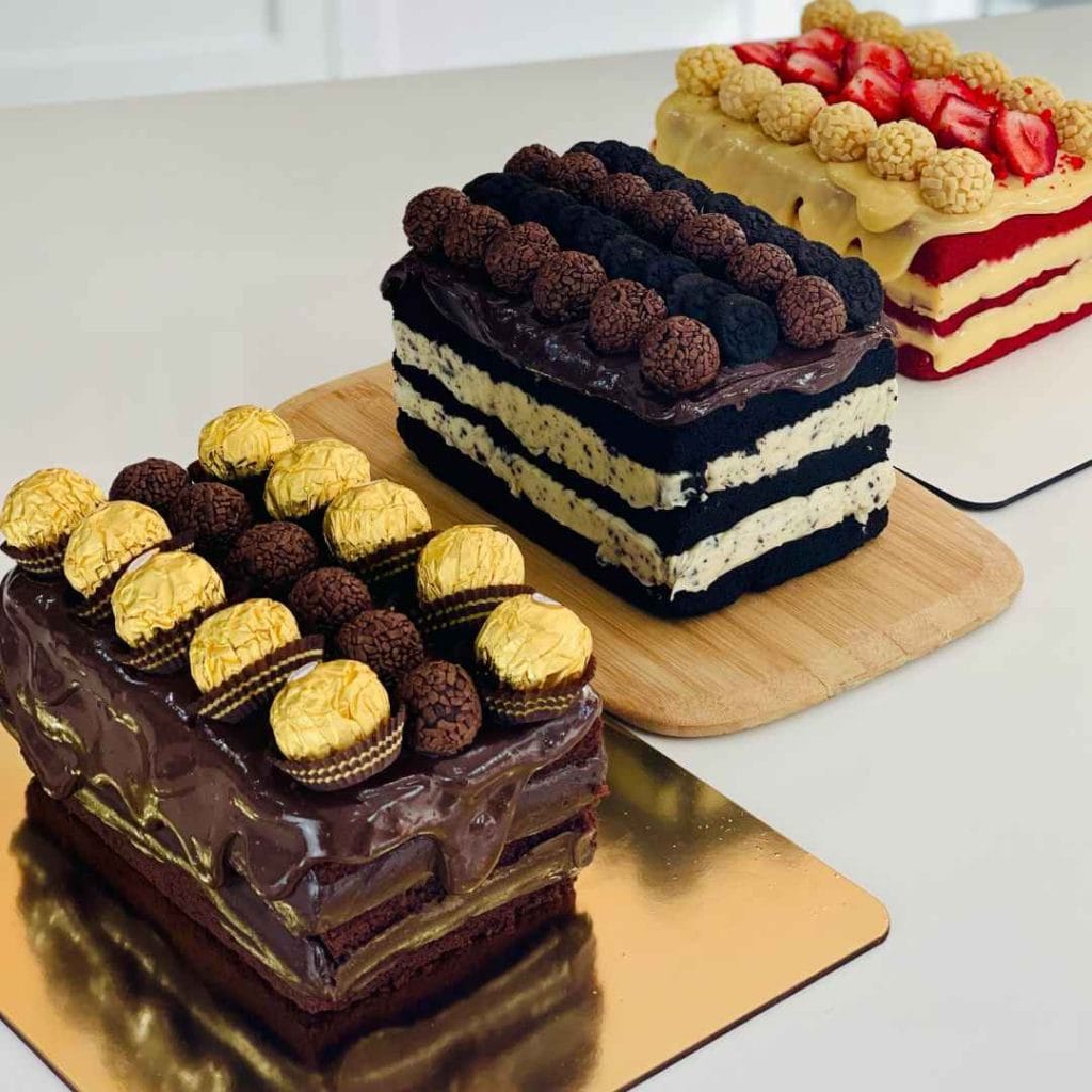 Três bolos slice cakes retangulares com recheios em camadas, decorados com Ferrero Rocher, brigadeiros e morangos, mostrando o acabamento de bolo de vitrine