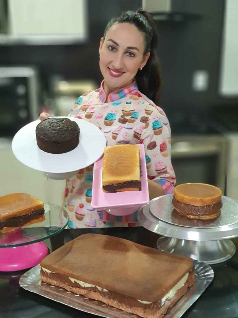 Chef confeiteira sorridente exibindo um pequeno bolo de chocolate e uma fatia de bolo recheada, demonstrando a montagem e o resultado das massas perfeitas