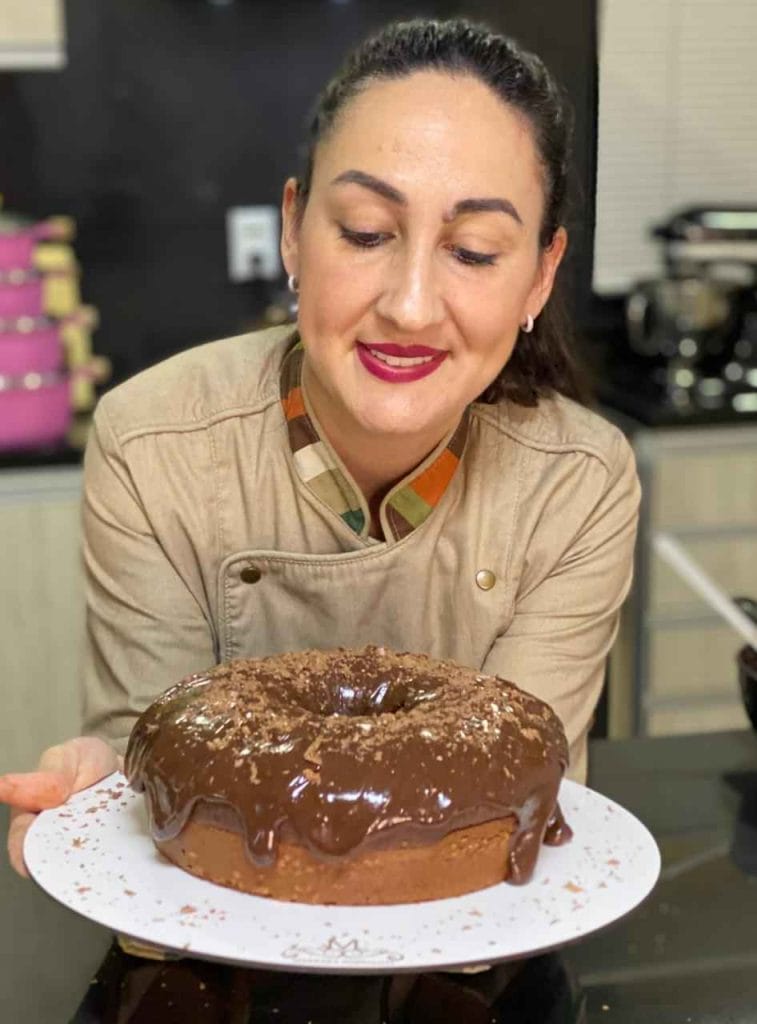 Chef confeiteira sorrindo com um bolo fit (sem glúten ou açúcar) de chocolate com cobertura brilhante e raspas no topo, provando que bolos fit vendem