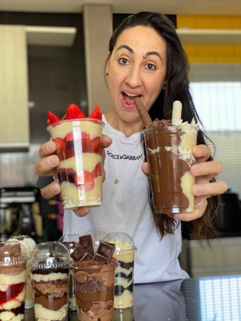 Chef confeiteira sorridente exibindo sobremesas no copo, incluindo copo de morango com creme branco e copo de chocolate com bolacha, conforme receitas de doces para vender
