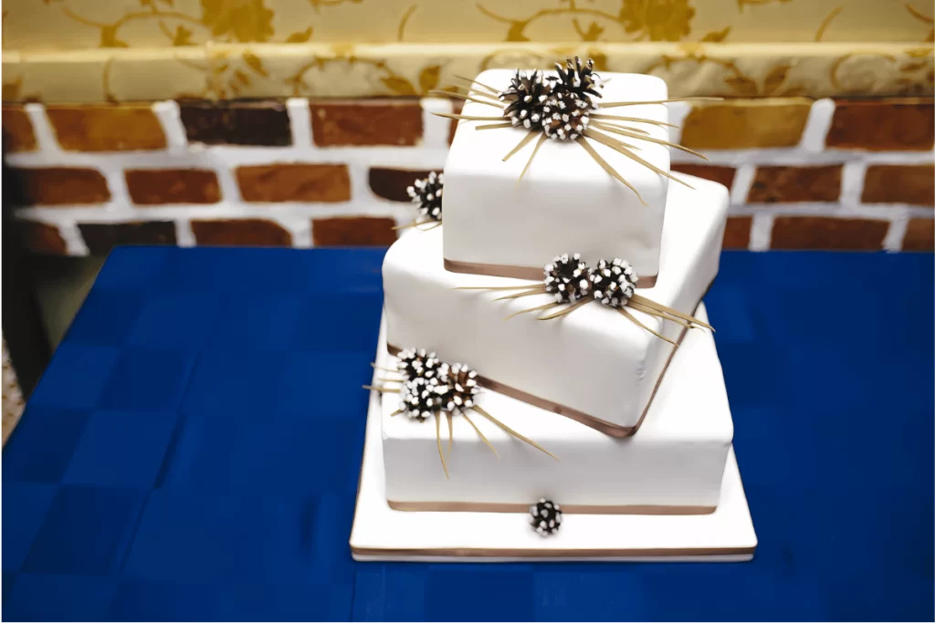 Bolo de casamento cake design quadrado branco de 3 andares, decorado com pinhas e fitas marrons, ilustrando a precisão da engenharia de bolos e design geométrico
