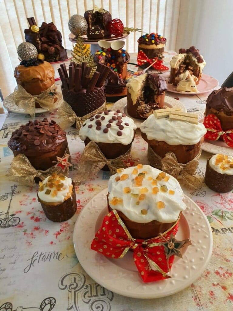 Mesa farta com diversos panetones e chocotones decorados, de diferentes tamanhos e sabores, prontos para a venda. Ilustra a variedade de um cardápio profissional e o potencial de lucro para confeiteiras.