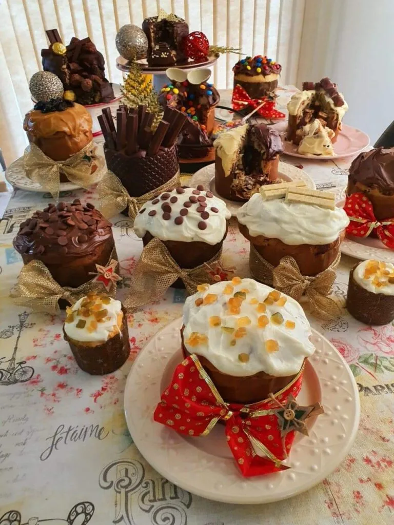 Mesa farta com diversos panetones e chocotones decorados, de diferentes tamanhos e sabores, prontos para a venda. Ilustra a variedade de um cardápio profissional e o potencial de lucro para confeiteiras.