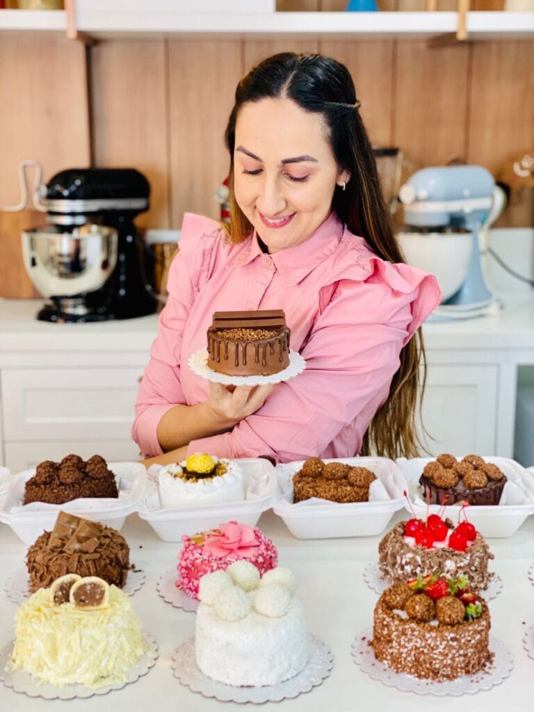 Chef confeiteira em camisa rosa apresentando uma mesa com vários minibolos de chocolate, coco, morango e kit kat, perfeitos para a apostila bento cakes e minibolos