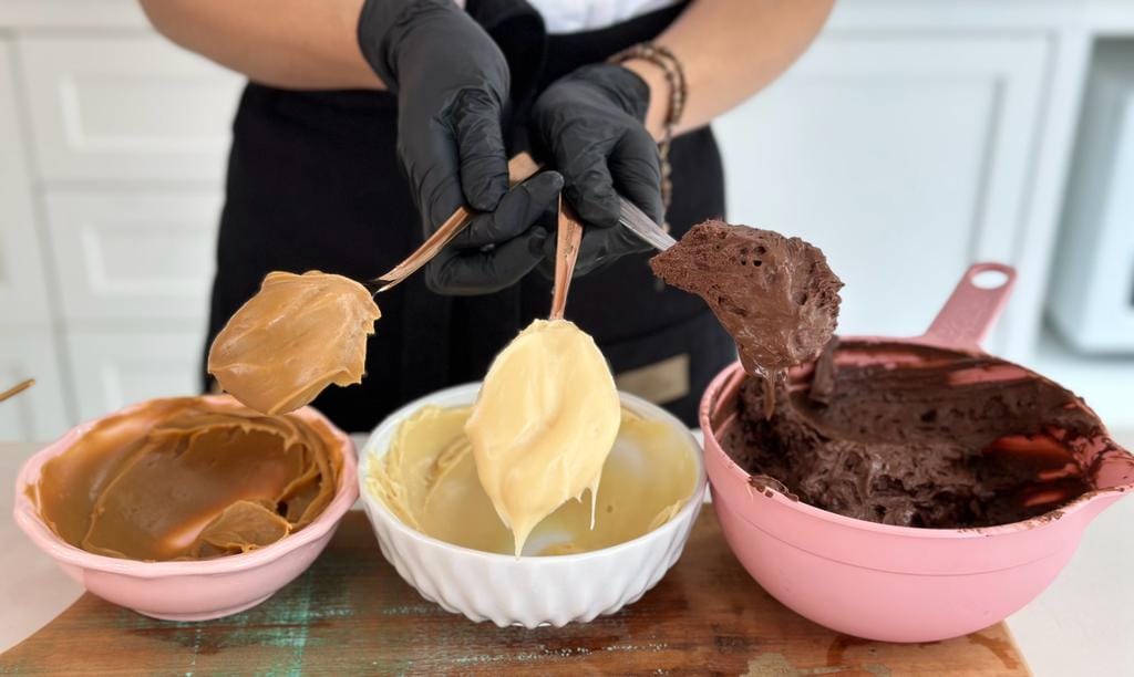 Mãos com luvas mostrando colheres com recheios cremosos de doce de leite, creme branco e chocolate, essenciais para bolos e doces de uma apostila de recheios