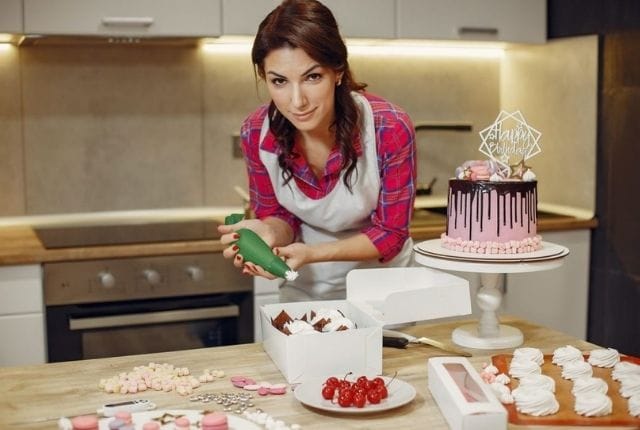 Confeiteira sorrindo em sua cozinha, decorando doces ao lado de um bolo de aniversário drip cake e embalagens, simbolizando o sucesso e a escala do negócio.