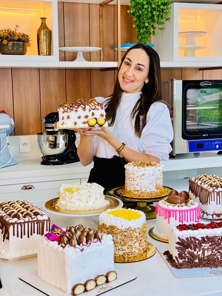 Confeiteira sorridente segurando um bolo de vitrine e rodeada por outros bolos de festa e bolos quadrados, com decorações ricas e coberturas brilhantes