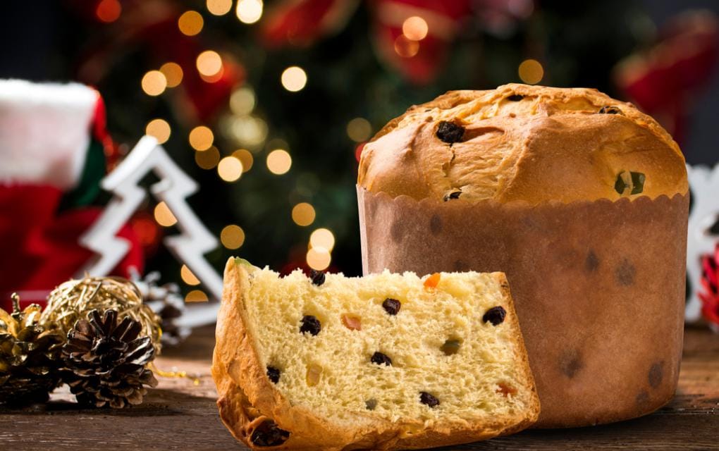 Foto de um Panetone tradicional com frutas cristalizadas ou gotas, em um cenário de Natal com luzes e enfeites. Esta imagem evoca o sentimento natalino para campanhas de marketing digital.